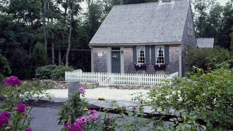Cape Cod-style house with a shingled roof behind a white picket fence