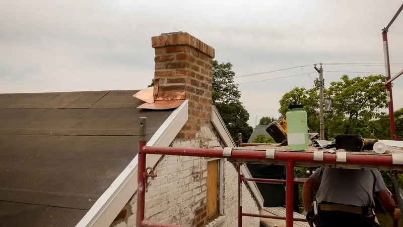 Mason working beside a brick chimney during roof work, with flashing at the base